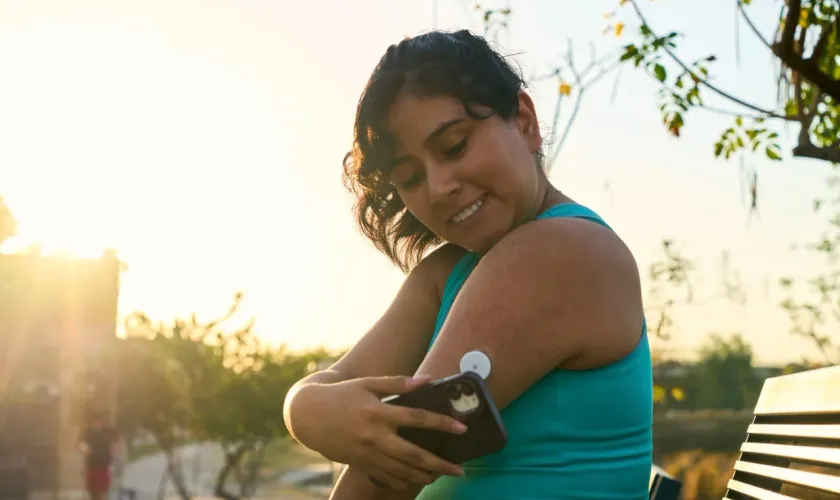 A woman checking her continuous glucose monitor with her phone while outdoors at sunset